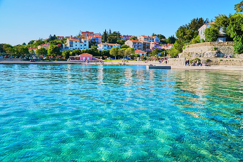 A photorealistic image of people enjoying various water sports activities near Liburnian Sun condominiums. Focus on the pristine beaches, clear water, and sunny weather. The camera angle is a wide shot, capturing the entire scene. Include elements like boats, kayaks, and paddleboards. 4k resolution.